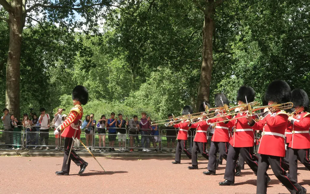 British guards in red uniforms and bearskin hats marching and playing trombones during a parade with spectators behind a fence.