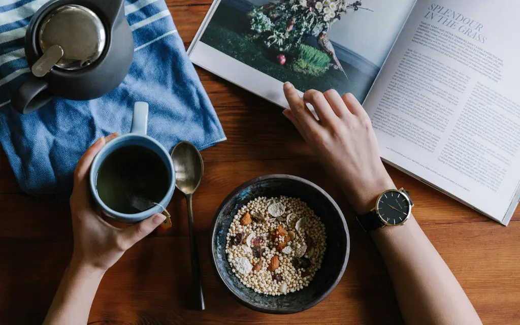 Person holding a blue cup of tea near a bowl of cereal, metal spoon, teapot on a blue cloth, and an open magazine on a wooden table.