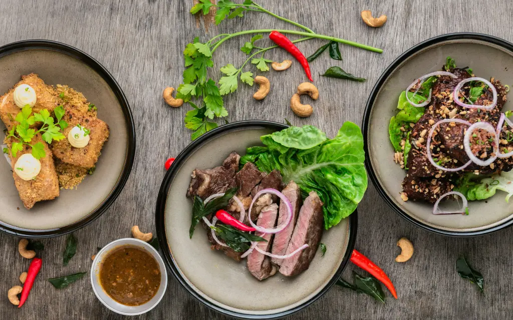 Three plates of food arranged on a wooden surface featuring breaded fried items with dollops of sauce, sliced steak garnished with red chili and onions on lettuce, and grilled meat topped with onion rings and crushed nuts, surrounded by cashew nuts, red chilies, and fresh herbs.