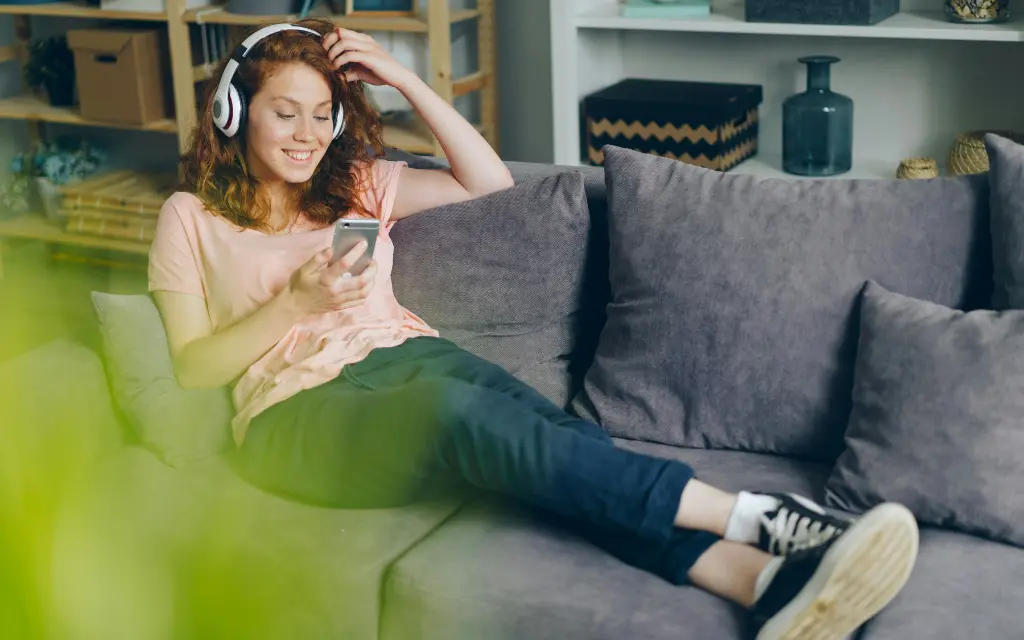 Young woman with headphones sitting on a gray couch, smiling while using her smartphone.