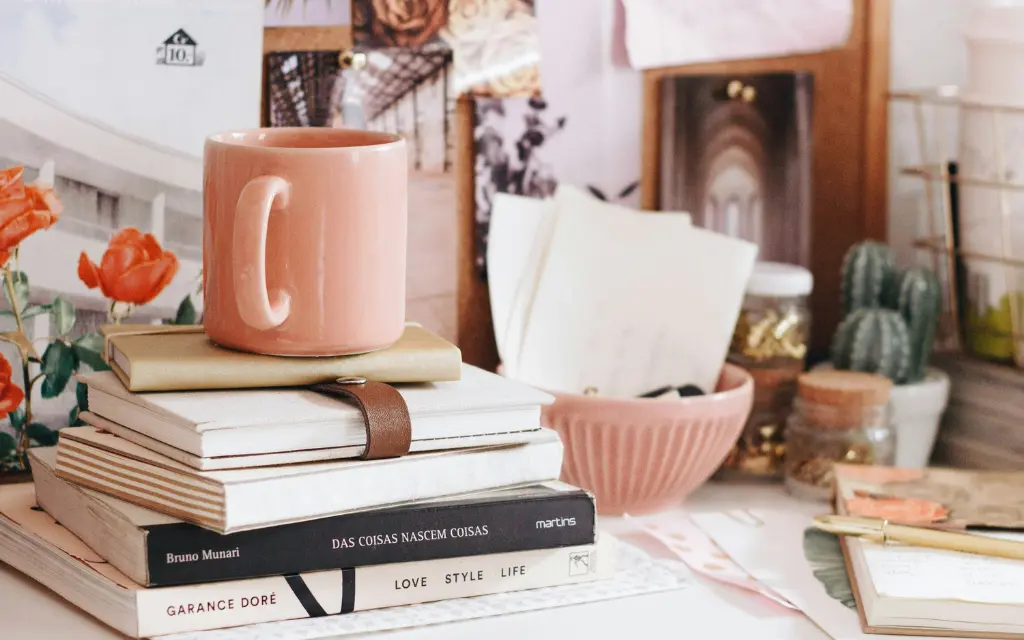Pink ceramic mug on top of a stack of books and notebooks with flowers and desk accessories in the background.
