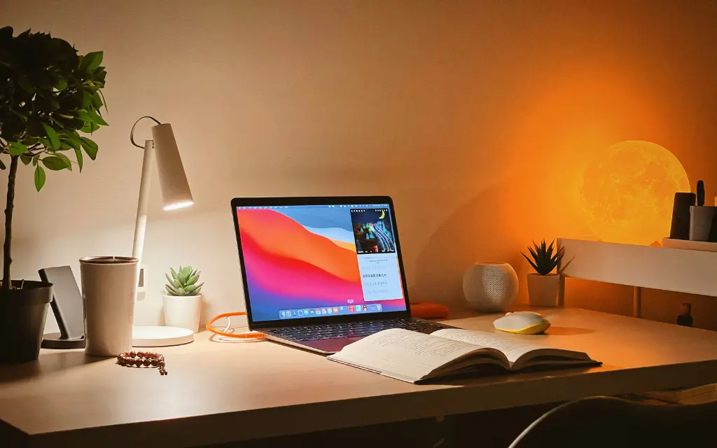 A cozy desk setup with a laptop, open book, desk lamp, potted plants, and a warm glowing moon-shaped light.